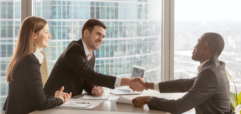 executive and job candidate shaking hands across desk, outside the window, an office tower gleams in the sun